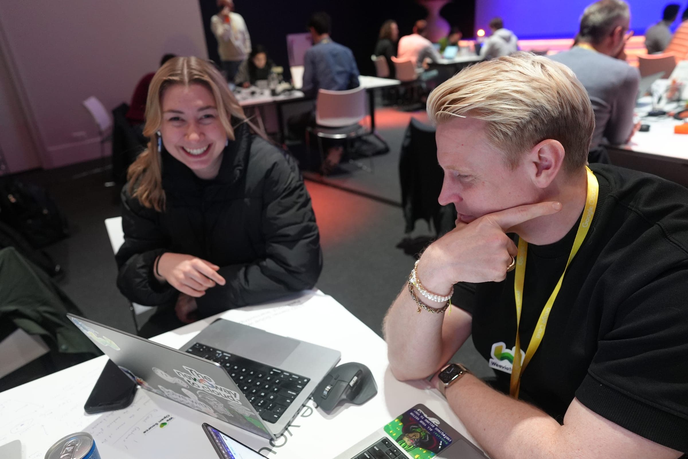 Victoria and a colleague at a hackathon table, laptops open, both grinning at something off-camera.