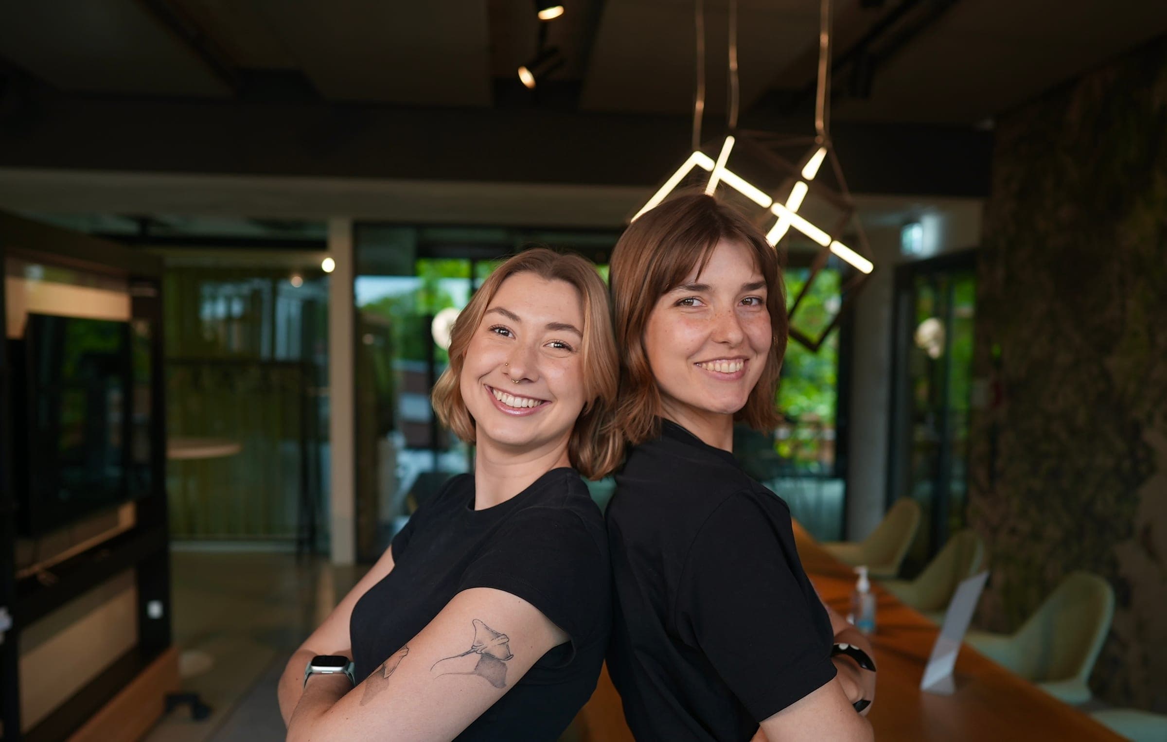 Victoria and a friend standing back to back in a bright modern room, arms crossed, both in black.