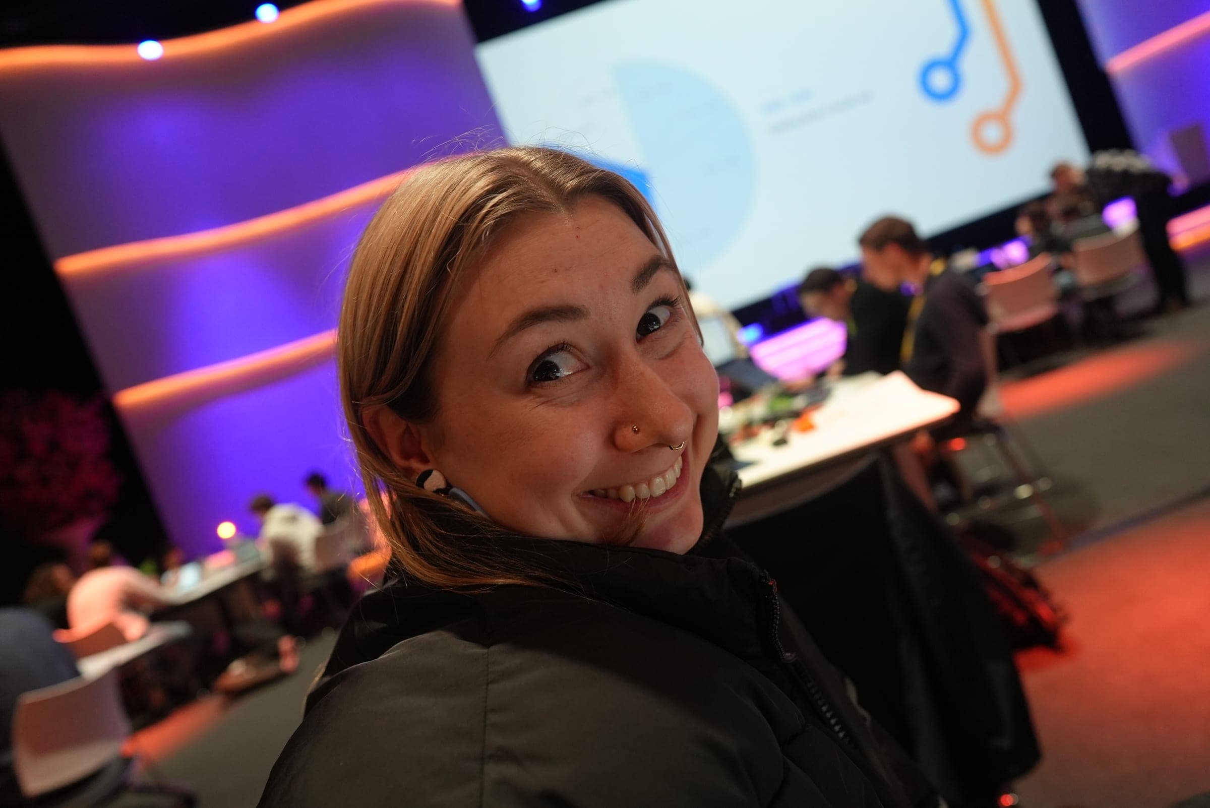 Victoria turning to the camera at a conference, purple stage lighting and a presentation screen behind her.