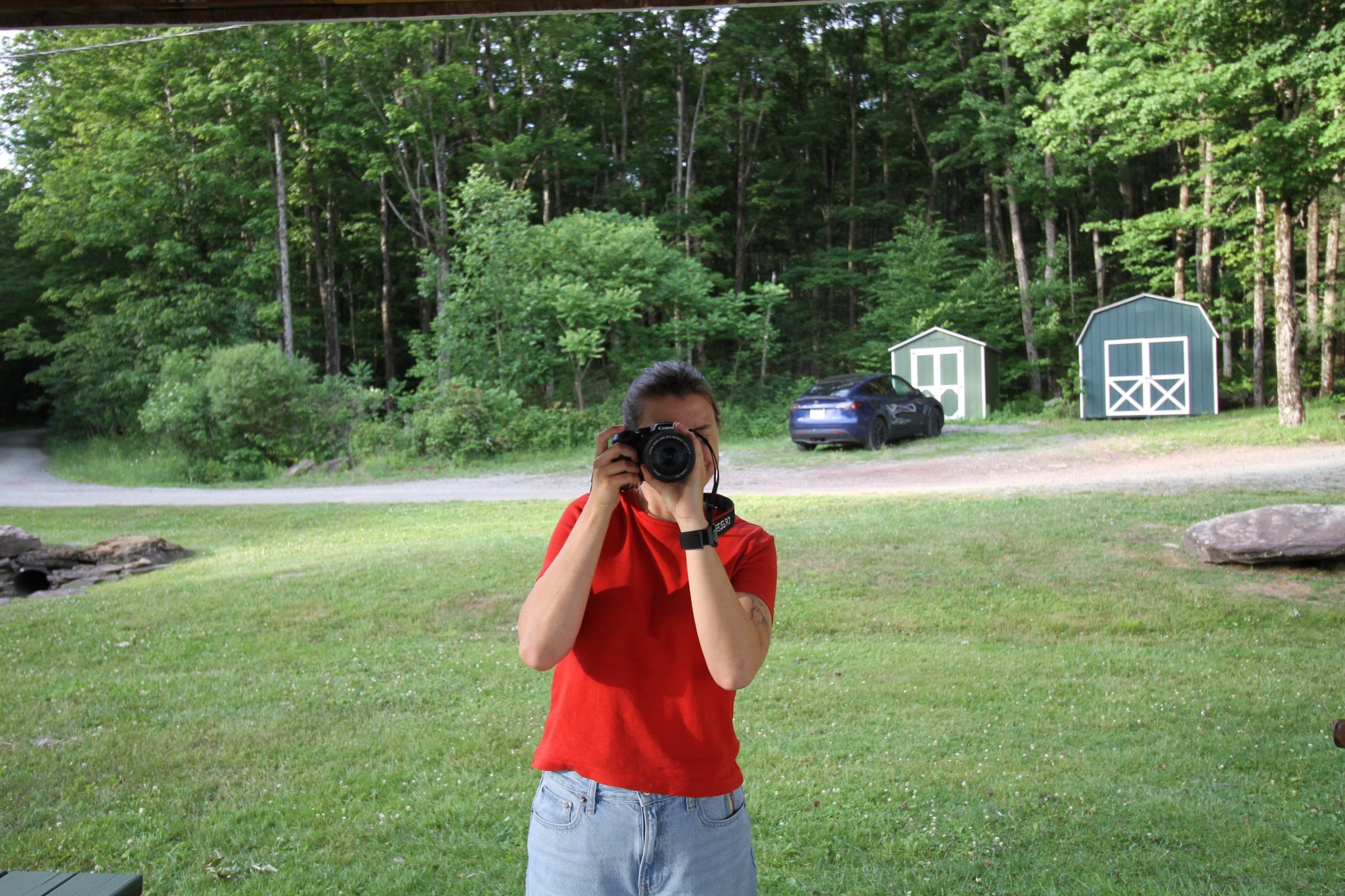 Victoria in a red T-shirt raising a camera to her eye on a wide green lawn.