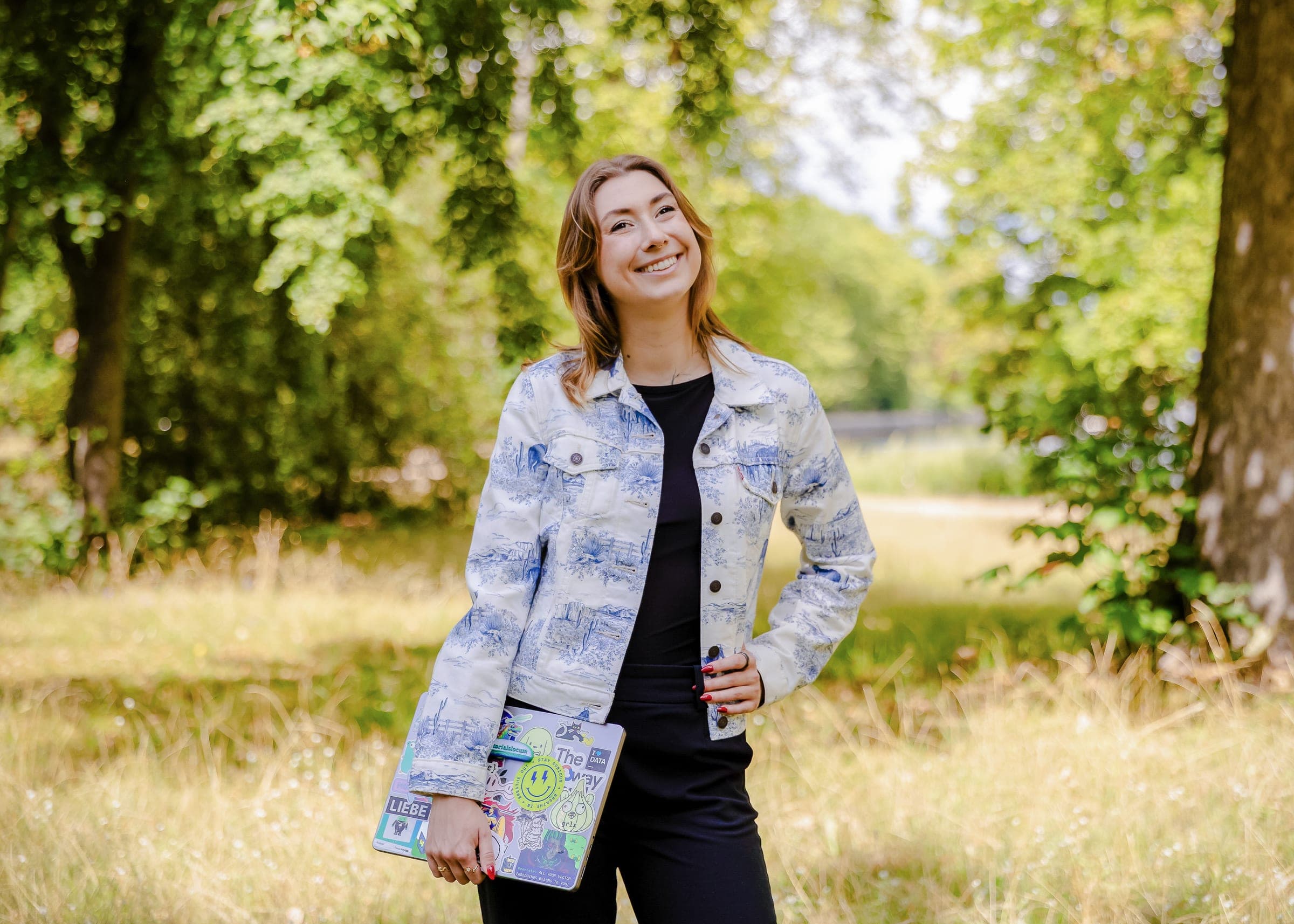 Victoria laughing in a sunlit meadow, holding a sticker-covered laptop, wearing a blue toile denim jacket.